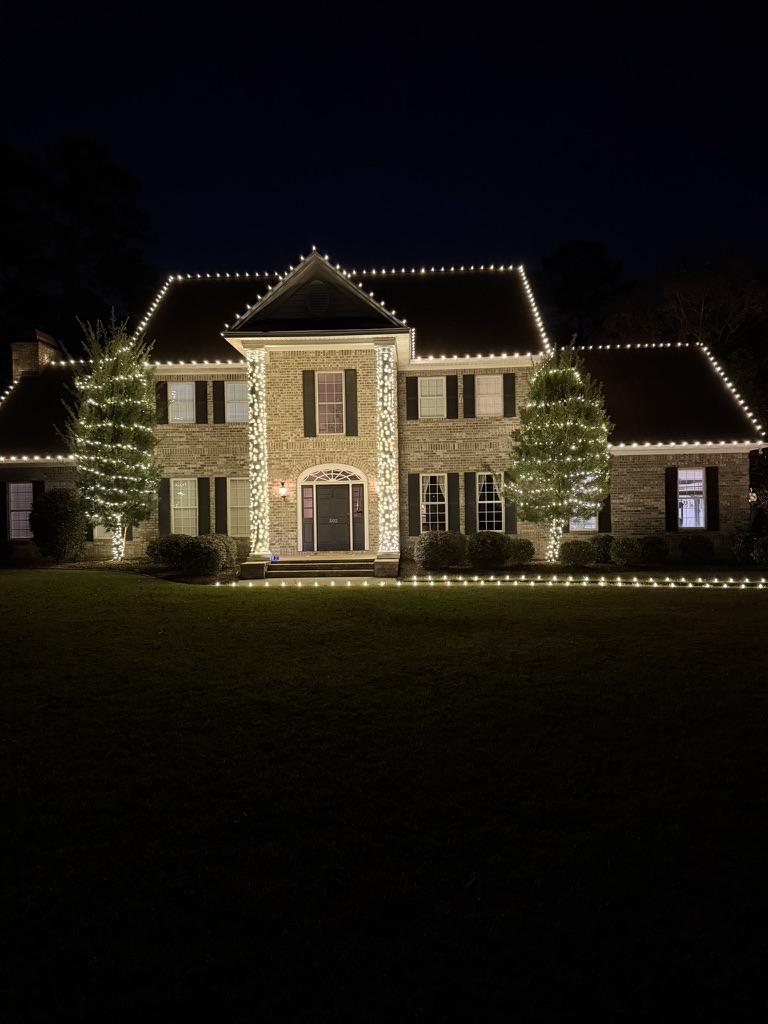 Large brick Savannah-area home with warm white roofline lighting, wrapped trees, and lit pathway